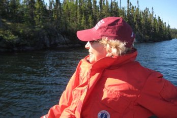 Photo of the late Dr. Sheila Seaman, illuminated by sunlight and sitting reclined in a boat in a lake in the middle of a Canadian boreal forest, facing towards viewer but looking left, wearing sunglasses and field jacket / PFD. She is smiling.