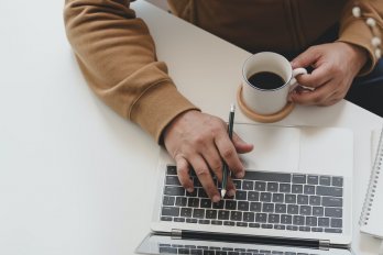 Hands and forearms of person using notebook computer at white table while drinking coffee and holding a pen.