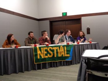 6 students seated, panel style, at table in hotel conference room, facing camera, with large banner displaying letters 'NESTVAL' draped over front of table.