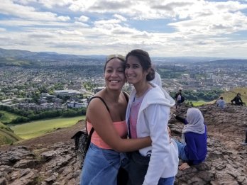 Raquel Bryant and Julie Beck facing camera in half-embrace atop rock outcrop in front of green pastoral scenery surrounding Edinburgh