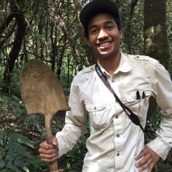 Portrait of Dr. Justin Richardson in field clothing in a forest, smiling at camera, holding shoveled with one hand, blade up.