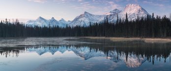 Placid Alaskan lake reflecting stunning scene of boreal forest with sweeping snow-capped mountain peaks in background with a blue sky nearing the golden hour prior to sunset.