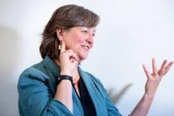 Picturre of Dr. Michele Cooke in front of white wall, gesturing to right of viewer, fingers splayed towards ceiling,and pointing at her ear.