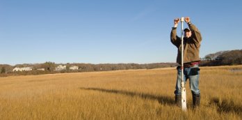 A grassy marsh in autumn duringa blue-sky day, where a male-presenting scientist in workcoat and jeans, is driving a soil-coring device, that is taller than them, into the ground