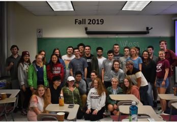 Over a dozen members of the U-Mass Geography club posing for a group photo within a classroom.