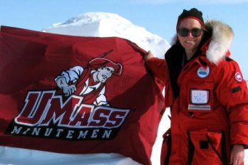 Photo of Dr. Julie Brigham Grette, smiling, wearing arctic cold-weather gear and sunglasses, holding U-Mass Minutemen flag in front of mountainous, snow-covered arctic landscape