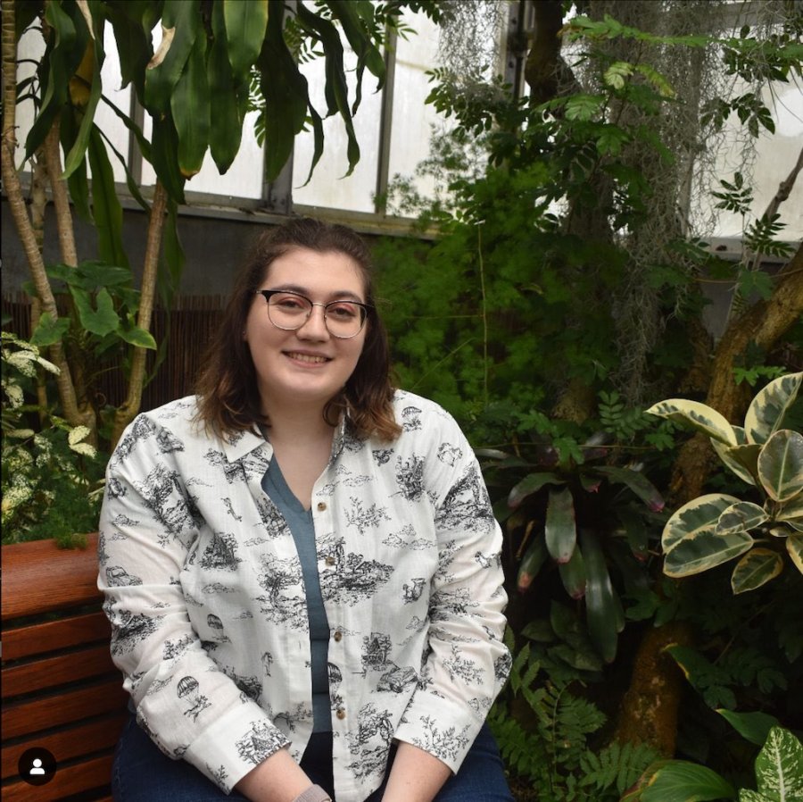 Helen Sajo, sitting on a wooden bench inside a verdant greenhouse.